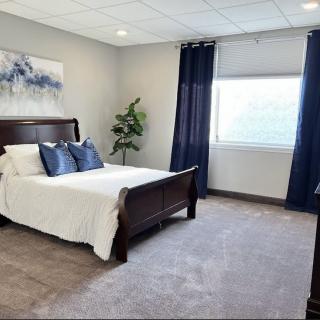 A well-decorated bedroom with a wooden bed frame, white bedding, and blue accent pillows. A large window with dark curtains allows natural light to fill the space.