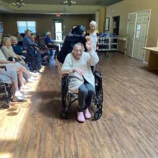A joyful moment in a care facility as an elderly resident waves from her wheelchair, surrounded by others enjoying their time.