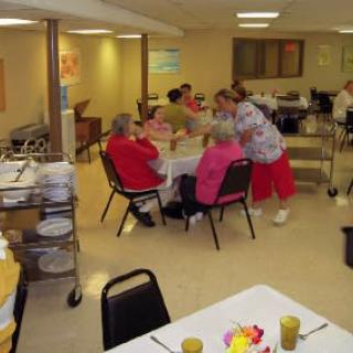 A group of seniors gather around a table in a dining area of a care facility.