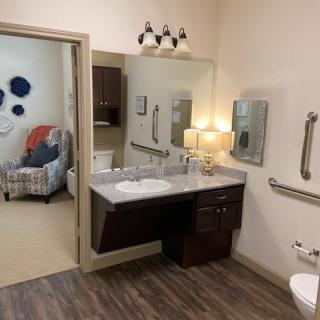 A spacious and accessible bathroom with a sink, mirror, lamp, and grab bars. The room features wood-style flooring and is connected to a cozy sitting area.
