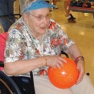 An elderly woman in a wheelchair holding an orange bowling ball indoors.