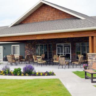 A patio with chairs and tables outside a building, surrounded by landscaping and a well-maintained lawn.