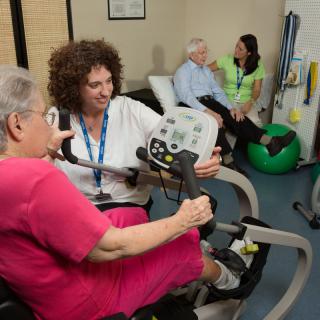 A healthcare professional assists a senior woman with exercise equipment in a therapy session, while another pair engages in conversation in the background.