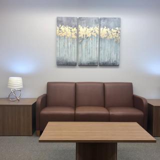 A modern office waiting area featuring a brown sofa, wooden table, lamps, and abstract artwork on the wall.