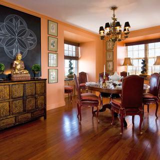 An elegant dining room with warm tones, featuring a wooden table, upholstered chairs, and a prominent chandelier. The room is tastefully decorated with artwork and a Buddha statue on a side cabinet.