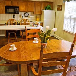 A cozy kitchen with a wooden dining table and chairs, featuring a refrigerator, microwave, and decorative flowers on the table.