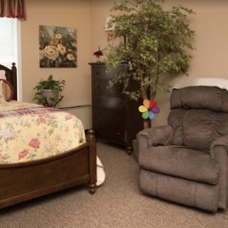 A cozy bedroom featuring a floral bedspread, a comfortable recliner, a dresser, and decorative elements including a potted plant.