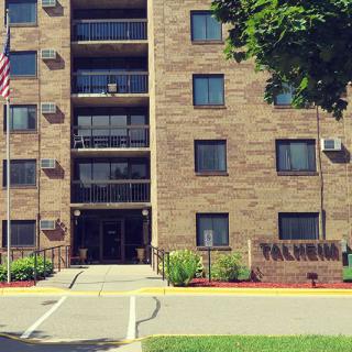 A brick apartment building with an entrance, surrounded by greenery and a flagpole in an urban setting.