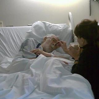 A person visits an elderly patient lying in a hospital bed.