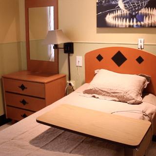 A hospital room with a bed, overbed table, nightstand, mirror, and wall art.
