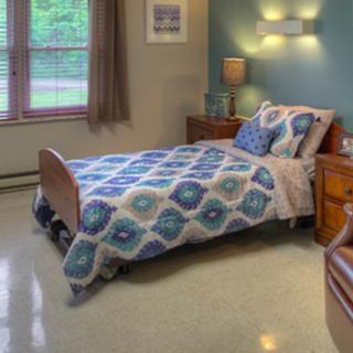 A cozy bedroom with a single bed featuring a patterned bedspread, a nightstand with a lamp, and a large window.