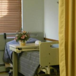 A hospital room with a bed, a vase of flowers on the table, and a curtain partially drawn.