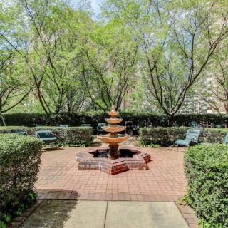 A tranquil garden with a central tiered fountain surrounded by lush trees and benches.