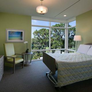 A hospital room with a large window offering a view of greenery outside, featuring a single bed, chair, and modern interior decor.