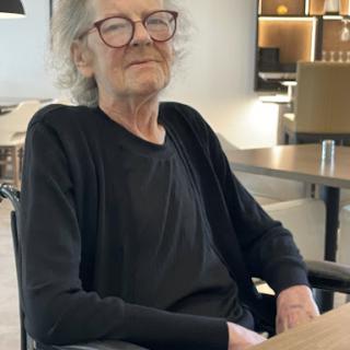A relaxed elderly person sitting indoors, wearing glasses and a black cardigan.