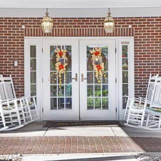 A welcoming entrance with double doors adorned with wreaths, flanked by white rocking chairs on a brick porch.
