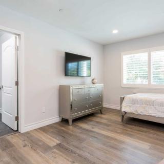A modern bedroom with minimalist decor featuring a bed, dresser, mounted TV, and a window with shutters.