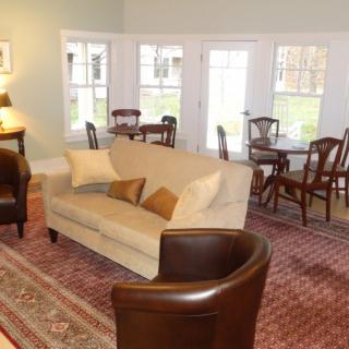A cozy living room featuring a beige sofa, brown armchairs, and a dining area with wooden chairs beside large windows.