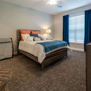 A modern bedroom with a neatly made bed, a lamp on the side, and blue curtains draping over a window.