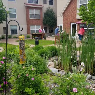 A beautifully maintained garden area with lush greenery and a small pond, set against the backdrop of an apartment complex. People are enjoying the serene environment.