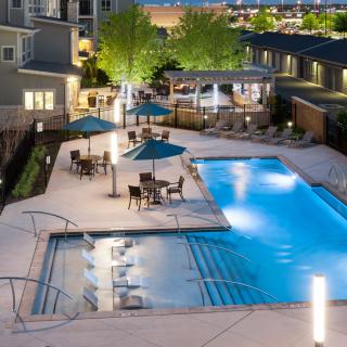 An inviting outdoor swimming pool area in a residential complex, featuring patio furniture and umbrellas under garden lights during the evening.