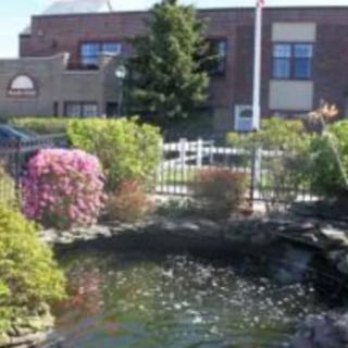 A scenic view of a pond surrounded by flowers and bushes, with a fence and building in the background.