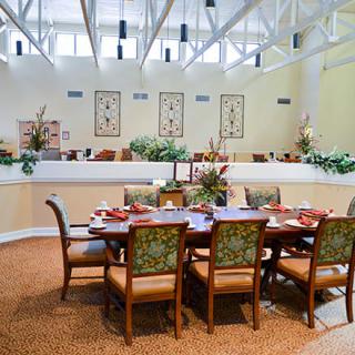 A modern dining room with a large table set for a meal, surrounded by chairs and decorated with plants and artwork.