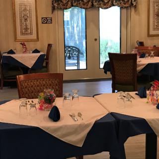 A cozy restaurant interior featuring elegantly set tables with blue and white tablecloths, decorated with flowers and a view of the outdoors.