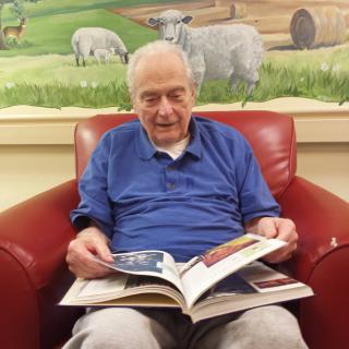 An elderly man is sitting in a red armchair, reading a book with a painting of sheep in the background.