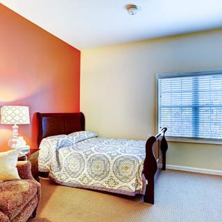 A cozy bedroom featuring a bed with patterned bedding, an upholstered chair, a lamp on a side table, and a window with blinds.