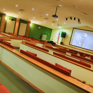 An empty lecture hall with rows of desks facing a large projector screen.