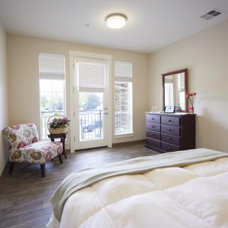 A cozy bedroom with natural light, featuring a bed, dresser, and armchair.