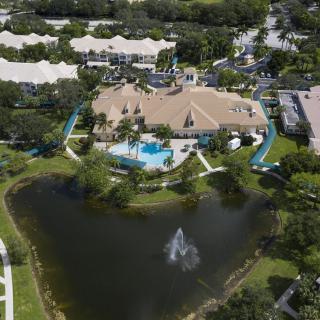 Aerial view of a residential complex featuring a large swimming pool, landscaped areas, and a nearby lake with a fountain.