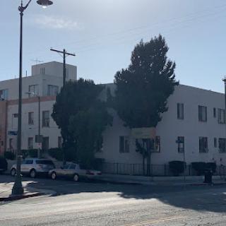 A view of a street scene featuring a building, trees, and parked vehicles under clear skies.