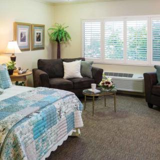 A cozy bedroom interior featuring a bed with a quilted cover, a sofa and armchairs, and a window with blinds.