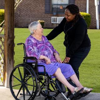 A caregiver interacts with an elderly woman in a wheelchair outdoors.