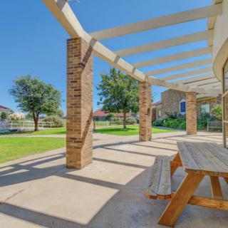 A serene outdoor patio area with a wooden table, surrounded by greenery and sunlight.