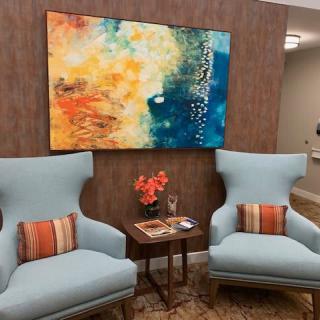 A cozy lounge area with two blue armchairs, a small table with magazines and flowers, and a colorful abstract painting on the wall.