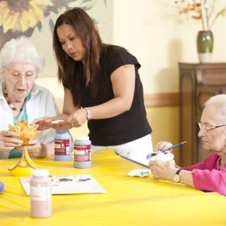 An art therapy session at a senior center where elderly individuals engage in painting activities with the help of a caregiver.