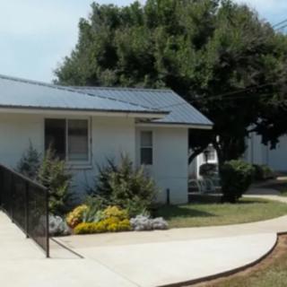 A small house with solar panels on the roof, surrounded by trees and landscaped plants along the driveway.