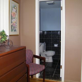 A bedroom with a wooden dresser and chair, leading to a small bathroom.