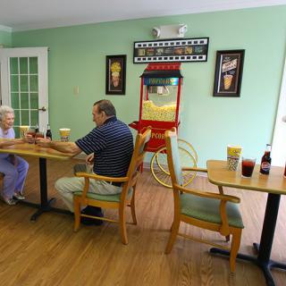 Elderly individuals enjoy a conversation in a cozy cafe setting with artwork and a popcorn machine in the background.