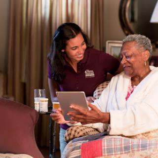 A caregiver assists an elderly woman with a tablet in a cozy home setting.