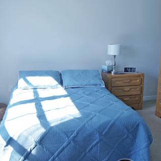 A serene bedroom featuring a blue bedspread and a nightstand with a lamp, illuminated by soft natural light.