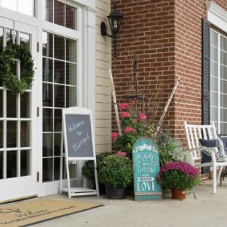 A charming storefront with a chalkboard sign, surrounded by flowers and garden tools, in front of a brick building.