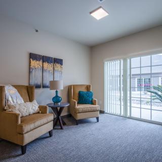 A cozy living room with two armchairs, a side table with a lamp, artwork on the wall, and plenty of natural light streaming in through large windows.
