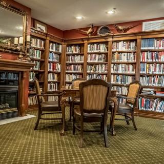 A cozy library with wooden bookshelves, a central table surrounded by chairs, and a fireplace.