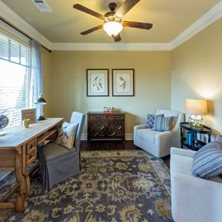 A cozy living room with elegant furniture, featuring a desk and comfortable armchairs, enhanced by a decorative rug and artwork on the walls.