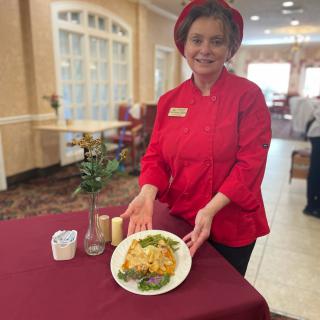 A chef in a red uniform proudly presents a plated dish in a restaurant setting.