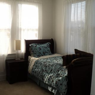 A cozy bedroom with a single bed by the window, featuring patterned bedding and a nightstand with a lamp.